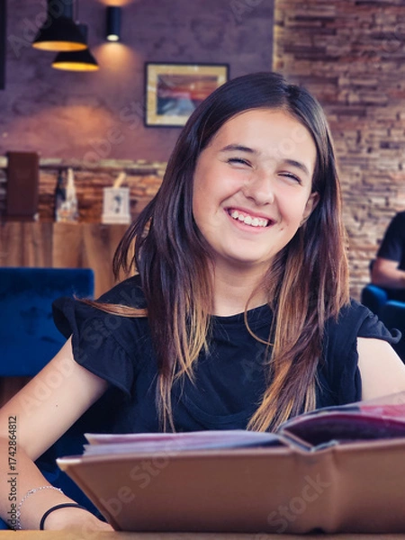 Fototapeta Smiling teenage girl sitting in a cozy cafe, holding a menu and enjoying a relaxed moment indoors.