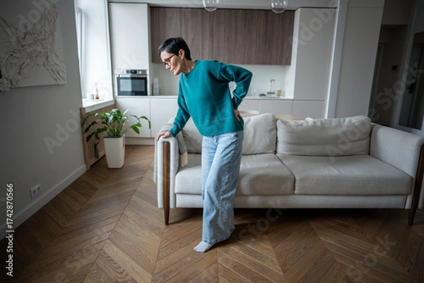 Fototapeta Struggling middle aged woman twisted in pain trying to rise from couch due to back issue. Herniated disc, chronic joint disease flare up, lumbar strain, movement restriction, spinal discomfort