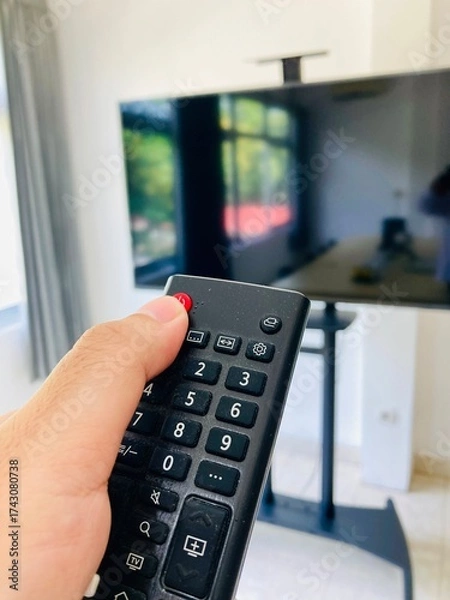 Fototapeta Close-up of a person’s hand holding a TV remote control while pointing it at a flat-screen television in a bright living room