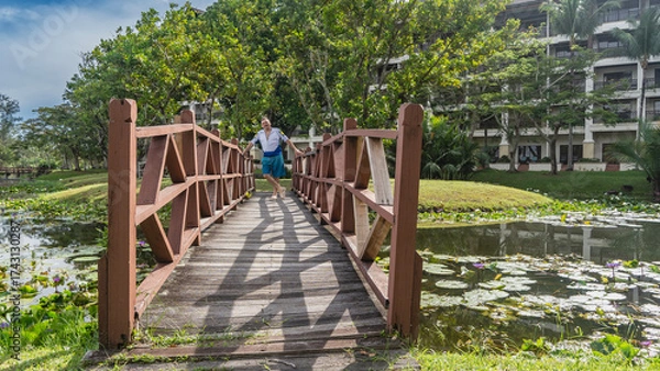 Fototapeta A man stands on a wooden pedestrian bridge over a pond, smiling, holding onto the railing. Leaves, flowers of water lilies on the water surface. Tropical trees, the hotel building in the background. 