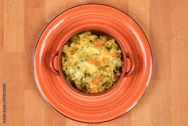 Fototapeta A portion of baked Brussels sprouts and potato with melted cheese in a small ceramic cocotte, served on a rustic orange plate on a wooden surface