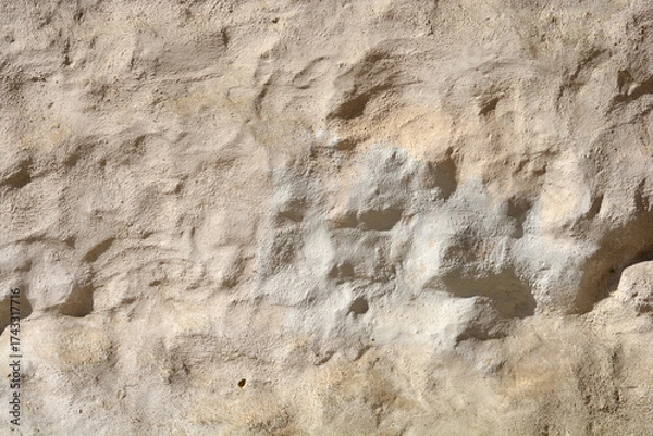 Fototapeta A close-up horizontal shot of an extremely rough and uneven wall texture with thick, weathered light beige stucco under harsh sunlight