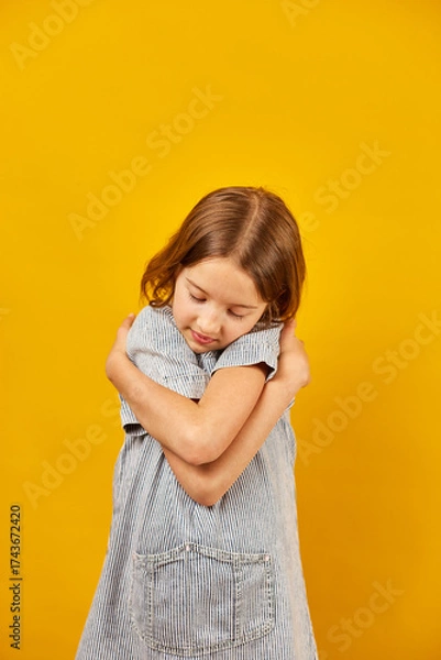 Fototapeta Young girl in striped shirt embraces herself against a bright yellow background while reflecting on her feelings