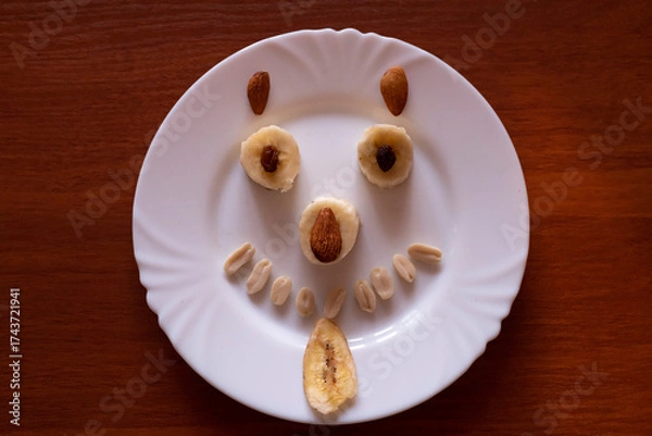 Fototapeta Creative fruit art featuring smiling face made of bananas and nuts on a white plate for playful snack time