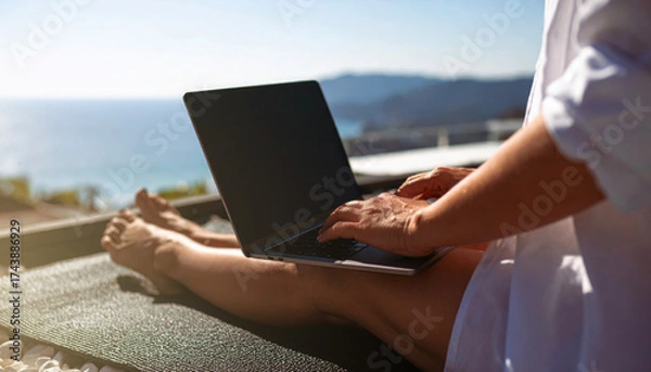 Obraz Faceles of a person sitting typing on a laptop while relaxing on a sunny balcony with a beautiful sea view.