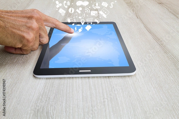 Fototapeta Man's hand touching on tablet computer with world map, social and business icons on wooden desk, business technology concept