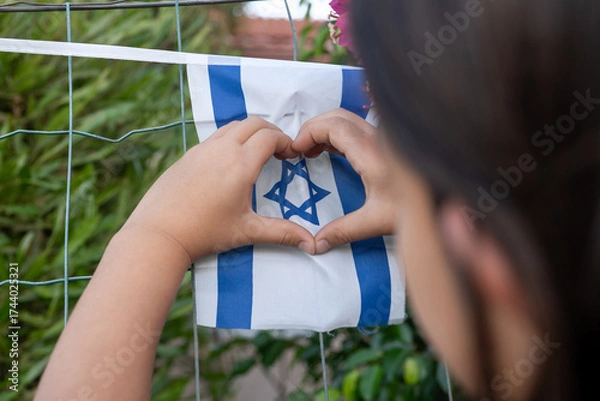 Fototapeta A child uses their hands to create a heart shape around an Israeli flag. The scene takes place outdoors, surrounded by green plants and bright flowers, conveying a message of love and patriotism.
