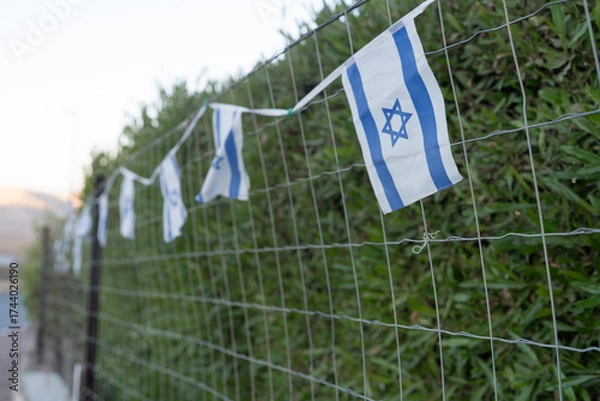 Fototapeta Small Israeli flags are attached to a wire fence, fluttering gently in the breeze. The surrounding greenery adds a natural touch to the scene, showcasing a peaceful atmosphere.