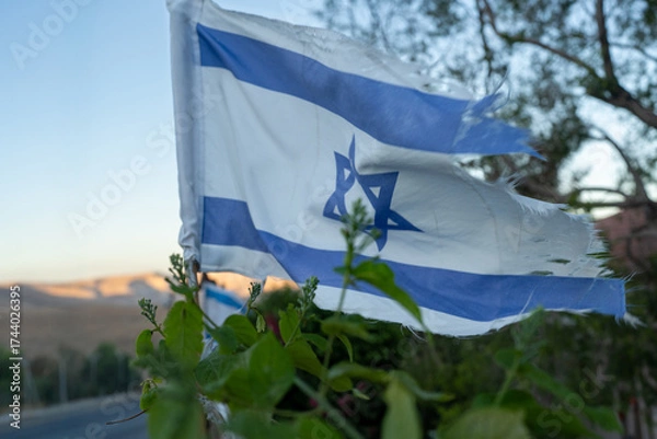 Fototapeta A frayed Israeli flag flutters gently in the breeze, surrounded by greenery. The sun sets behind distant hills, casting a warm glow on the landscape and flag, symbolizing resilience.