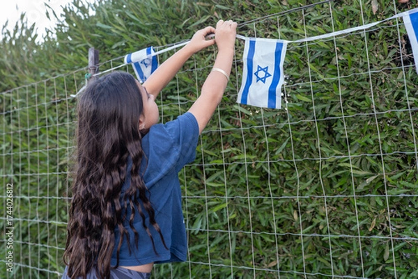 Fototapeta A young girl with long hair is carefully attaching small Israeli flags to a fence surrounded by greenery. The scene suggests preparations for a festive event.