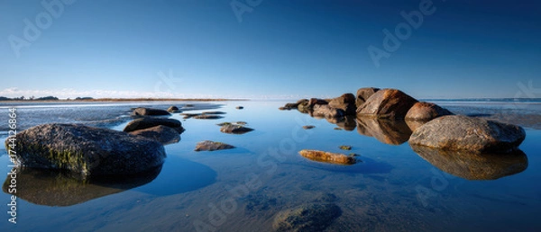 Fototapeta Calm ocean shore with rocks reflecting in water under clear blue sky