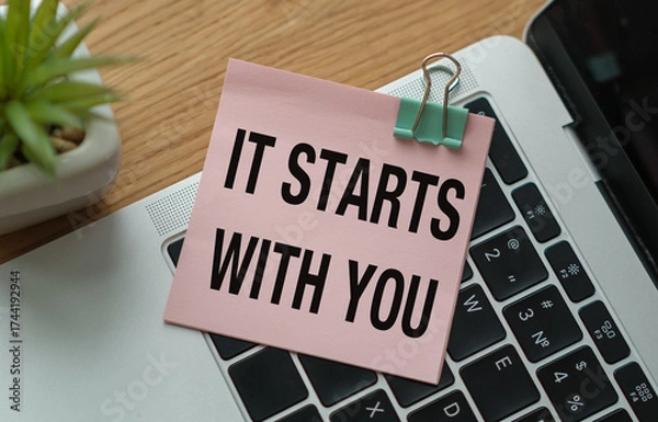 Fototapeta A pink piece of paper with the words It Starts With You written on it is sitting on top of a laptop keyboard