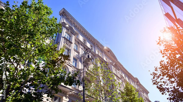 Fototapeta A residential building  by lush green trees and surrounded by  fresh vegetation on a clear sunny day. Modern architecture residential apartment building complex surrounded by greenery.