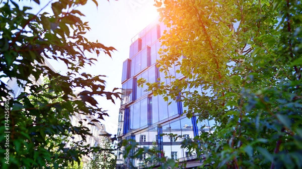 Fototapeta Glass office building surrounded with green trees.  Eco-friendly office building. Sustainable architecture of city reducing heat carbon dioxide modern city environment.