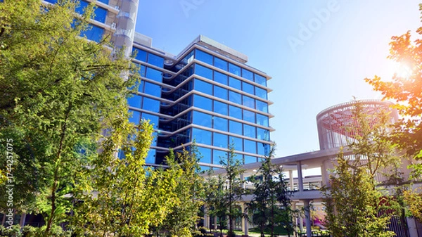 Fototapeta Glass office building surrounded with green trees.  Eco-friendly office building. Sustainable architecture of city reducing heat carbon dioxide modern city environment.