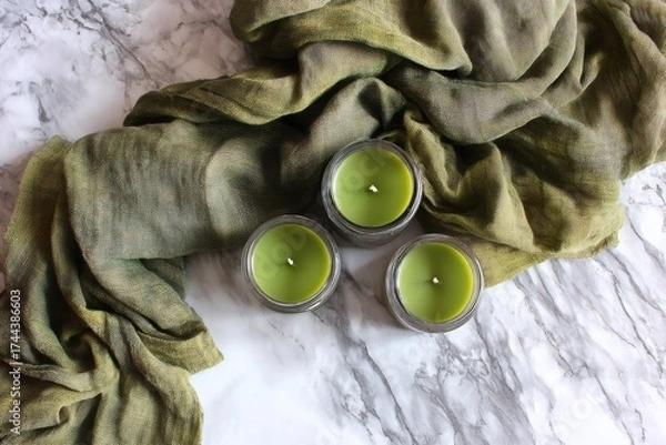 Fototapeta Three green scented candles in glass jars with a draped fabric on white marble, top view, calm atmosphere
