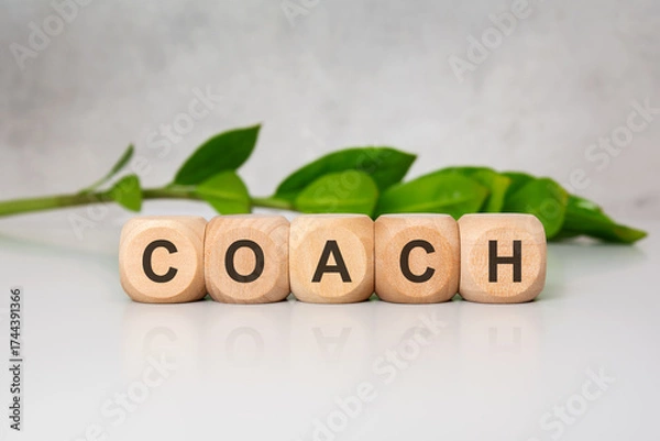 Fototapeta Wooden blocks spelling COACH on a white surface with greenery in background