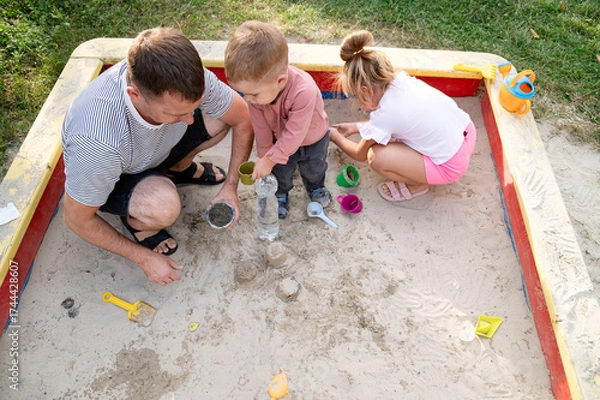Obraz Father enjoys playful moments with his children in a sandpit during a sunny afternoon in the park
