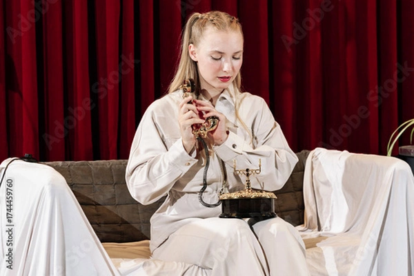 Fototapeta A young, beautiful actress sits on a sofa with an old telephone in her hand. Theater stage.