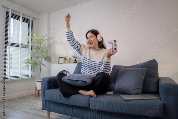 Fototapeta A young woman sits on a sofa wearing headphones while holding a game controller and playing a video game on her laptop. Her cheerful expression shows enjoyment and excitement.
