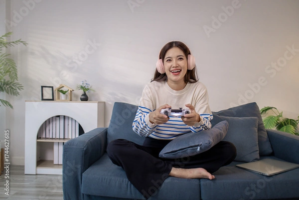 Fototapeta A young woman sits on a sofa wearing headphones while holding a game controller and playing a video game on her laptop. Her cheerful expression shows enjoyment and excitement.