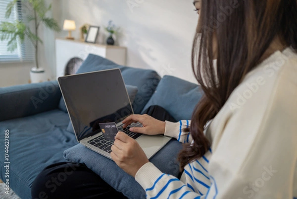 Fototapeta A young woman sits on a sofa using a laptop while holding credit cards, representing secure online payment and modern lifestyle. She smiles confidently while shopping from home.