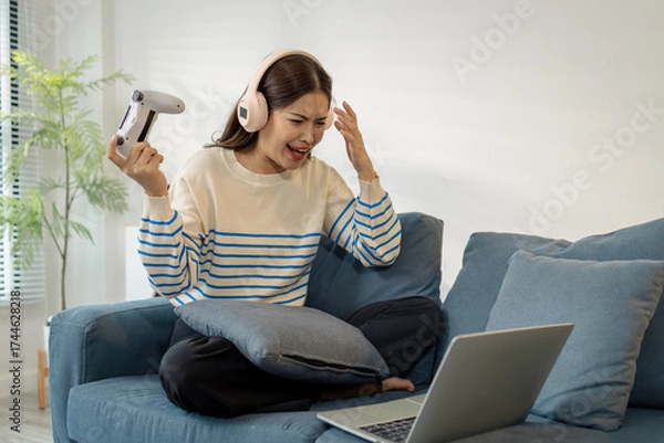 Fototapeta A young woman wearing headphones is playing a video game on her laptop, holding a controller with emotional expressions of joy, excitement, and frustration during gameplay.