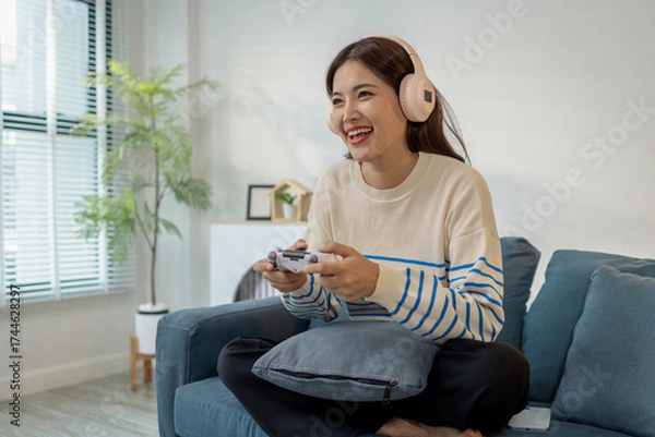 Fototapeta A young woman wearing headphones is sitting on a sofa while holding a game controller, showing joy, excitement, and focus as she plays a video game indoors.