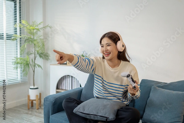 Fototapeta A young woman wearing headphones is sitting on a sofa while holding a game controller, showing joy, excitement, and focus as she plays a video game indoors.