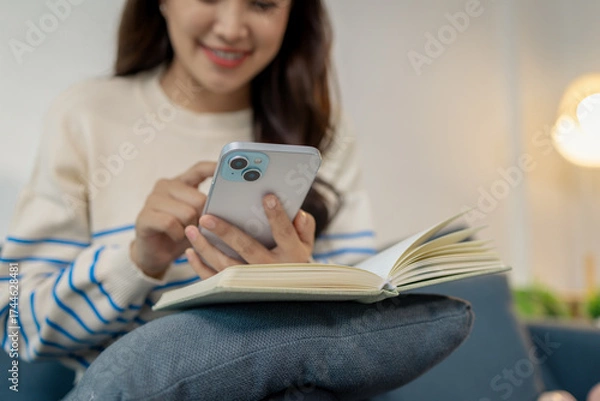 Fototapeta A young woman sitting comfortably on a sofa holding a smartphone while smiling and reading a message, with an open notebook resting on her lap in a bright cozy room.
