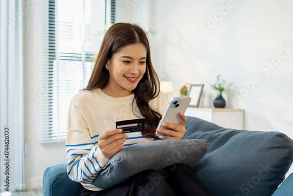 Fototapeta A young woman is sitting on a sofa holding a smartphone and credit card, smiling as she makes an online transaction. She looks relaxed and confident while shopping at home.
