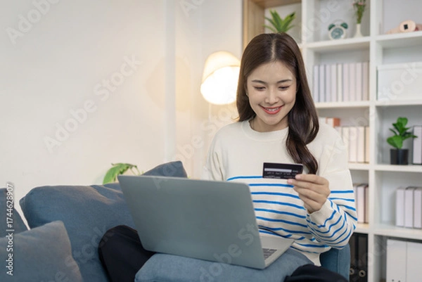 Fototapeta A young woman sits comfortably on a sofa using a laptop while holding a credit card, appearing to make an online payment. She smiles with satisfaction, reflecting ease and convenience.