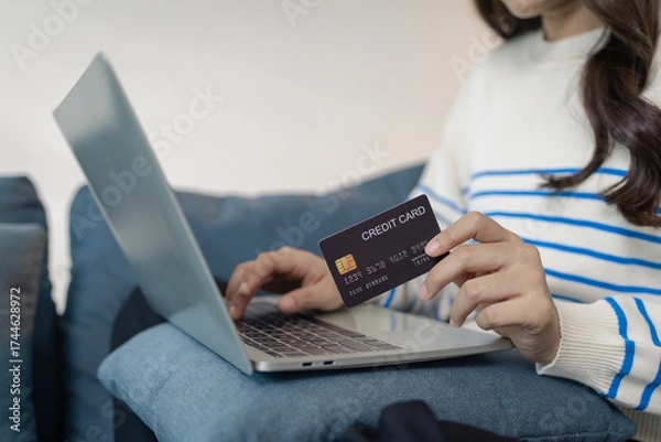 Fototapeta A young woman sits comfortably on a sofa using a laptop while holding a credit card, appearing to make an online payment. She smiles with satisfaction, reflecting ease and convenience.