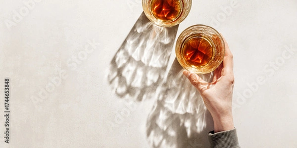 Fototapeta Alcohol Glasses with dark shadows at sunlight on beige table, hand holding glass, top view minimal style photo banner, aesthetic flat lay shiny glass at natural light, summer drink, copy space