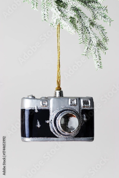 Fototapeta Minimal winter composition with toy camera Christmas ornament hanging from snowy fir branch on light gray background, creative holiday concept connecting photography, travel memory and New Year.