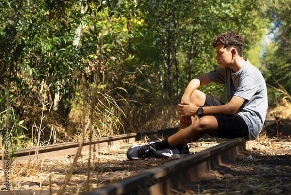 Fototapeta Young Boy Sits Thoughtfully on Abandoned Railway Tracks Surrounded by Overgrown Grass and Trees on a Sunny Day