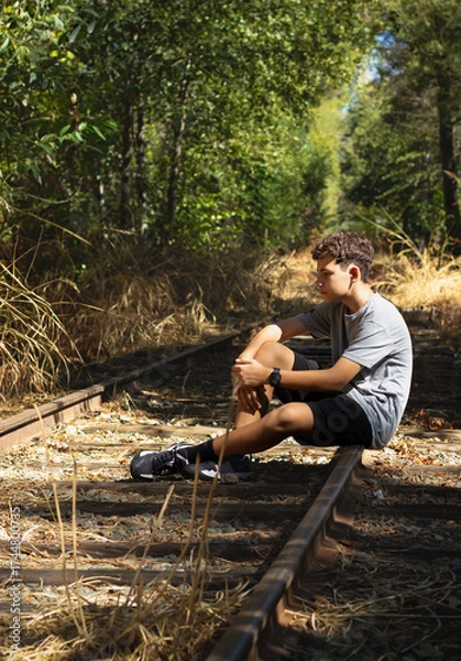 Fototapeta Boy Sitting on Train Tracks in a Forest During Daytime, Lost in Thought Among Tall Grass and Trees