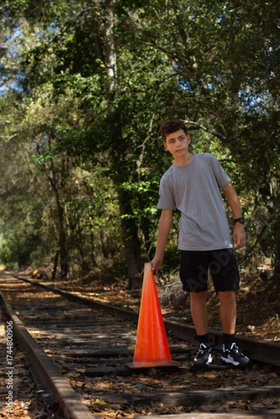 Fototapeta Young Boy Standing on Railway Tracks Holding an Orange Traffic Cone During a Sunny Afternoon in a Forested Area