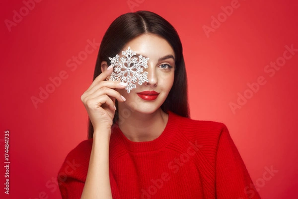 Fototapeta Beautiful young woman with red lipstick, shiny clean skin and face, showing glowing snowflake, near face and smiling, isolated on a red background. Christmas holidays concept