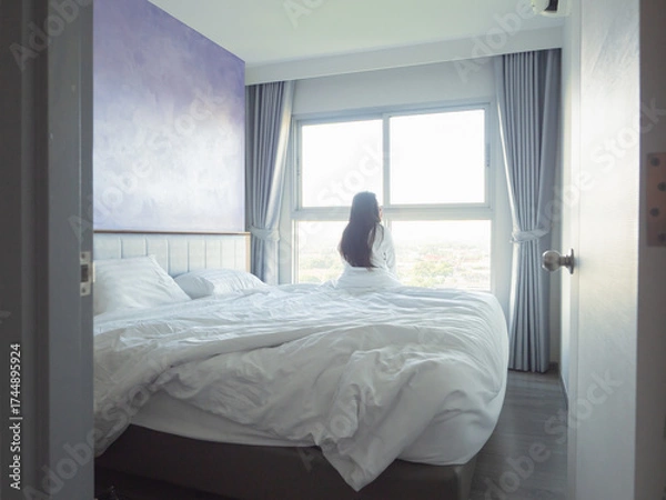Fototapeta Young woman relaxing in bed with white bedding, smiling in soft morning light by the window