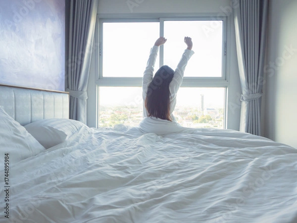 Fototapeta Young woman relaxing in bed with white bedding, smiling in soft morning light by the window