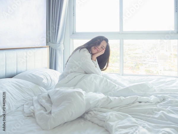 Obraz Young woman relaxing in bed with white bedding, smiling in soft morning light by the window