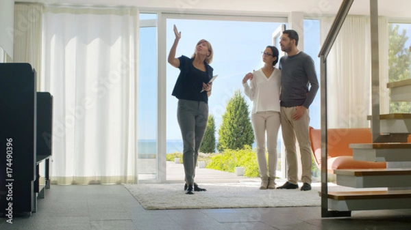 Fototapeta Professional Real Estate Agent Shows Stylish Modern House to a Beautiful Young Couple Who are in the Market for Purchasing/ Renting New Home. House Has Floor to Ceiling Windows and Seaside View.
