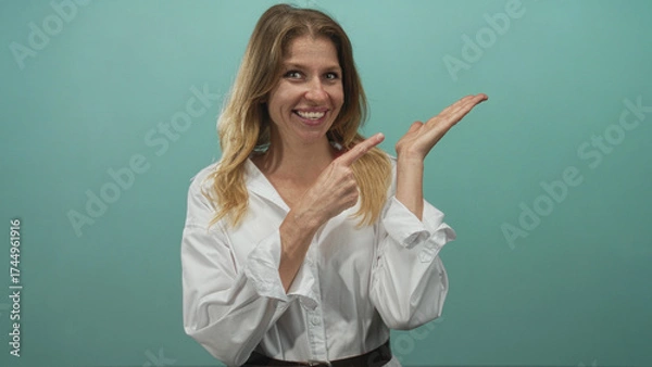Obraz Woman points finger to palm in studio teal backdrop wearing white shirt and belt; cheerful confidence.