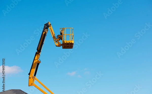Fototapeta Elevated boom lift platform cherry picker against clear blue sky during daytime works on construction site with bright sunlight illuminating the machinery