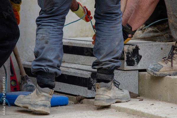 Fototapeta Team of builder workers lifting heavy concrete block for stairs construction. Teamwork and determination as a path to success concept.