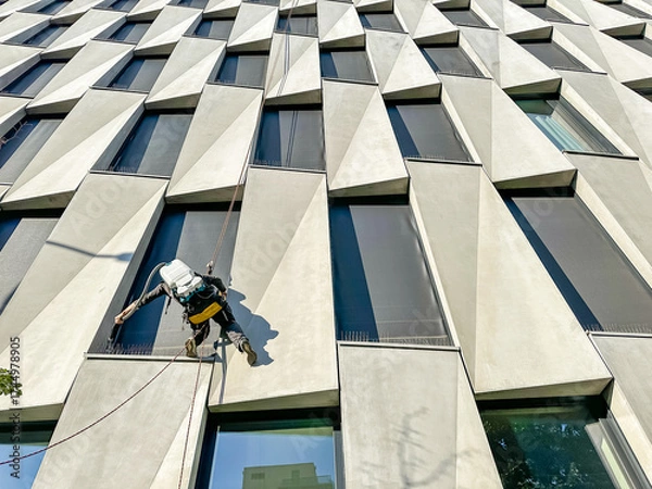 Fototapeta industrial climber, window cleaner works on office building at height of several floors on cables, washing windows of metropolis business center with mops and pressure water machines