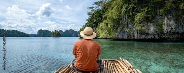 Fototapeta A serene view of a person sitting on a bamboo raft, gazing at lush green cliffs and calm waters under a blue sky, evoking tranquility and connection with nature.
