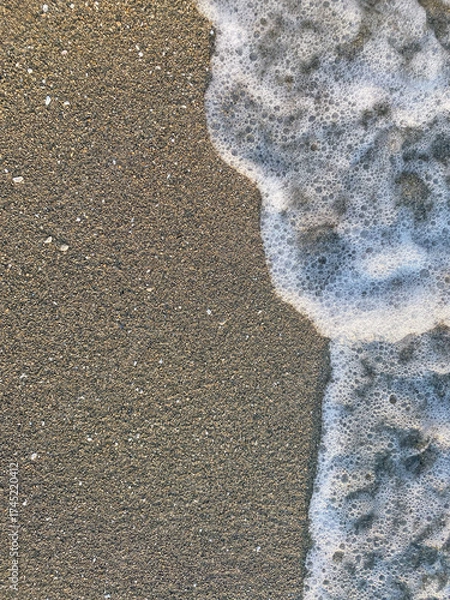 Fototapeta Waves gently wash over a sandy beach, leaving a trail of foam in the early morning light