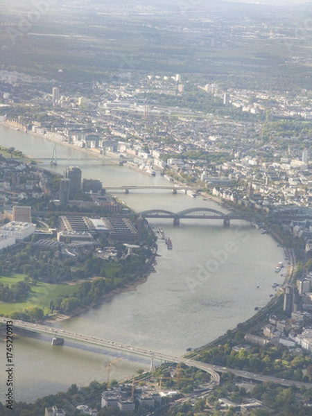 Fototapeta Aerial view of the Rhine River winding through Cologne city center, with bridges and green spaces under a cloudy sky